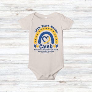 May include: A beige baby onesie with a blue and yellow rainbow design. The text "Little Heart Warrior" arches above the rainbow, with the name "Caleb" below. The phrase "I can do all things through Christ who gives me strength" is also printed on the onesie.