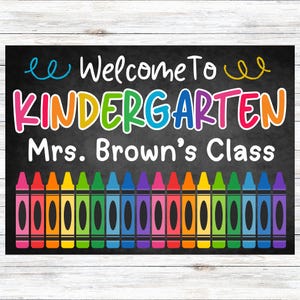 May include: A chalkboard-style sign with the words "Welcome To Kindergarten Mrs. Brown's Class" in colourful letters. A row of colourful crayons is at the bottom of the sign. The background is black with a white wooden frame.