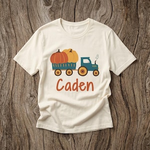 May include: A cream-coloured short-sleeve t-shirt featuring a graphic of a blue tractor pulling a trailer filled with orange and yellow pumpkins. The name Caden is printed in orange script lettering below the tractor illustration.