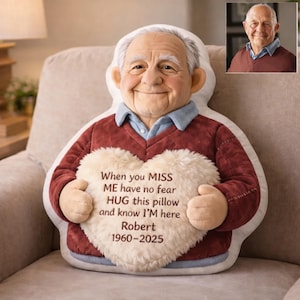 Puede incluir: Un cojín de felpa con forma de un anciano sonriente, con un suéter burdeos y una camisa con cuello azul claro. El cojín sostiene un corazón de felpa color crema con el texto "When you MISS ME have no fear HUG this pillow and know I'M here Robert 1960-2025."