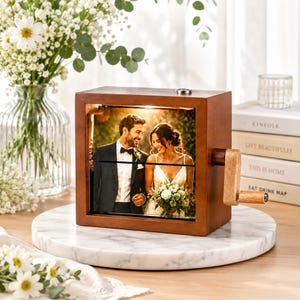 Op de afbeelding: Een bruine houten fotocube met een handslinger, met een trouwfoto. De kubus heeft een lichtgekleurde houten slinger. De foto toont een lachend stel. De kubus staat op een wit marmeren oppervlak, met bloemen op de achtergrond.