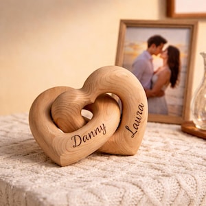 May include: Two interlocking wooden hearts, one engraved with "Danny" and the other with "Laura". A framed photo of a couple kissing is in the background. The hearts are on a textured white surface.
