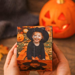 Op de afbeelding: Een foto kubus met Halloween-thema met een lachend kind in een heksen kostuum. De kubus toont een reeks afbeeldingen, met een uitgesneden pompoen op de achtergrond. De kubus wordt in iemands handen gehouden.