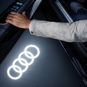Op de afbeelding: Een autodeurinterieur met een zwart paneel met raambediening en een lichtgekleurde mouw. Een helder wit Audi-logo wordt op de grond onder de open deur geprojecteerd, wat een gloeiend embleem met vier ringen op het donkere oppervlak creëert.