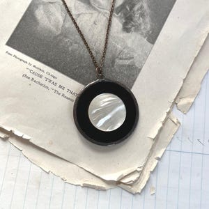 May include: A circular pendant necklace featuring a central iridescent white mother-of-pearl disc set within a smooth black circular frame. The pendant is suspended from a thin, dark metallic chain, creating a high-contrast, geometric design.
