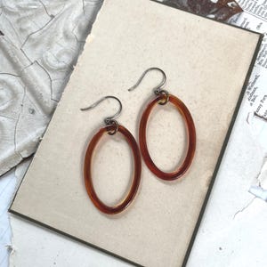 Puede incluir: Un par de pendientes ovalados con aros de resina translúcida de color ámbar suspendidos de ganchos metálicos oscuros. Los pendientes tienen un acabado liso y pulido con un diseño sencillo de aro abierto que cuelga verticalmente.