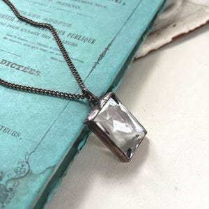 Puede incluir: Un collar con un colgante rectangular transparente y un marco plateado. La cadena oscura descansa sobre un libro azul vintage con texto en francés.