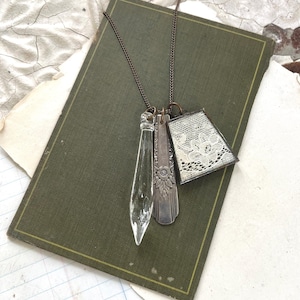 May include: A necklace featuring a thin bronze chain with three charms: a clear glass crystal pendant, a silver-toned metal piece with an embossed floral pattern, and a trapezoidal frame containing a piece of white lace fabric with a floral design.