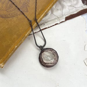 Puede incluir: Un collar artesanal con una cadena de metal oscuro y un colgante circular. El colgante presenta un centro texturizado plateado rodeado por un borde marrón rojizo oscuro. El collar se exhibe sobre un libro antiguo.