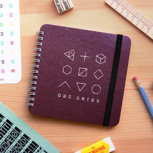 May include: A burgundy notebook with a spiral binding and a black elastic band closure. The notebook has a white graphic design of geometric shapes and the text "ODD GRIDS".