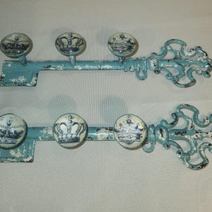 May include: Two decorative key-shaped wall hooks. The hooks are painted a distressed teal color with white accents. Each key has three round knobs with a blue and white crown design. The keys are made of metal.