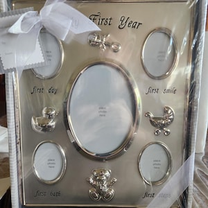 May include: Silver-toned baby photo frame with space for multiple photos. The frame has the words "First Year", "first day", "first smile", "first bath", and "first steps". Decorative elements include a baby carriage, teddy bear, and duck.