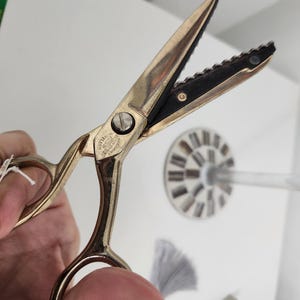 May include: Gold-colored fabric scissors with a black serrated blade. The scissors have a small inscription on the handle that reads "The Best Scissors Made In Germany".