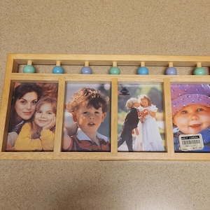 May include: A wooden picture frame with seven blue and green beads across the top. The frame holds five photos, including a family of three, a young boy, a couple, and a baby. The frame has a price tag attached to the bottom right corner.