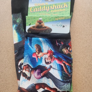 May include: A black golf towel featuring a colorful graphic print of characters from the movie Caddyshack. The towel includes a cardboard header card with the text Tribute to Caddyshack Collection, Hi Def Edge Towel, and David O'Keefe.
