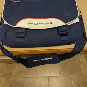 May include: A blue and white messenger bag with a red stripe and the text "BMW Sauber F1 Team" on the front. The bag has a shoulder strap and a zipper closure.