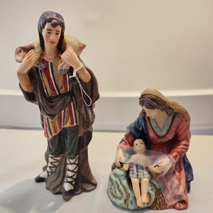 May include: Two ceramic figurines depicting a religious scene. One figure is a man in a brown robe carrying a lamb. The other is a woman in a red and blue robe holding a baby wrapped in a translucent cloth. The figurines are painted with detailed features.