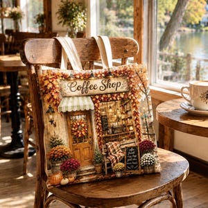 Op de afbeelding: Een canvas draagtas met een bedrukte illustratie van een koffiebar-gevel versierd met herfstbladeren, pompoenen en potbloemen. Het ontwerp bevat een houten deur, een gestreepte luifel en een klein krijtbordje met tekst.