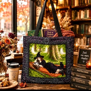 May include: A black tote bag featuring a purple ornate frame border and a central illustration of a witch in a black dress and hat lying on a blanket in a forest with a black cat, a broom, and three white ghosts in the background.