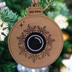 Może przedstawiać: Drewniana ozdoba choinkowa w kształcie kuli, z napisem "Elf Cam" na górze. Ozdoba posiada wzór płatka śniegu wokół czarnego okręgu z białą zakrzywioną linią i gwiazdą. Wisi na choince.
