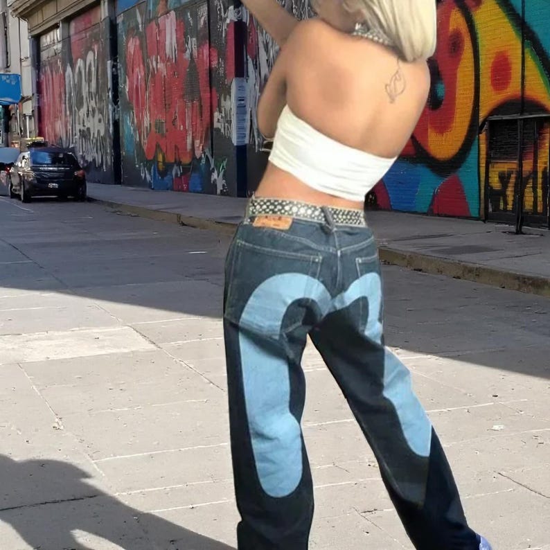 Puede incluir: Vaqueros de mezclilla de lavado oscuro con un dise&ntilde;o curvo azul claro en las piernas. Los vaqueros se combinan con un top de tubo blanco y un cintur&oacute;n estampado. El fondo presenta un mural de graffiti colorido.