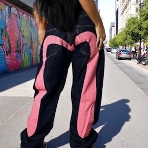 Puede incluir: Vaqueros de mezclilla azul oscuro con un gran dise&ntilde;o rosa en la parte trasera y los lados. Los vaqueros tienen un corte holgado. La foto est&aacute; tomada al aire libre en un d&iacute;a soleado.