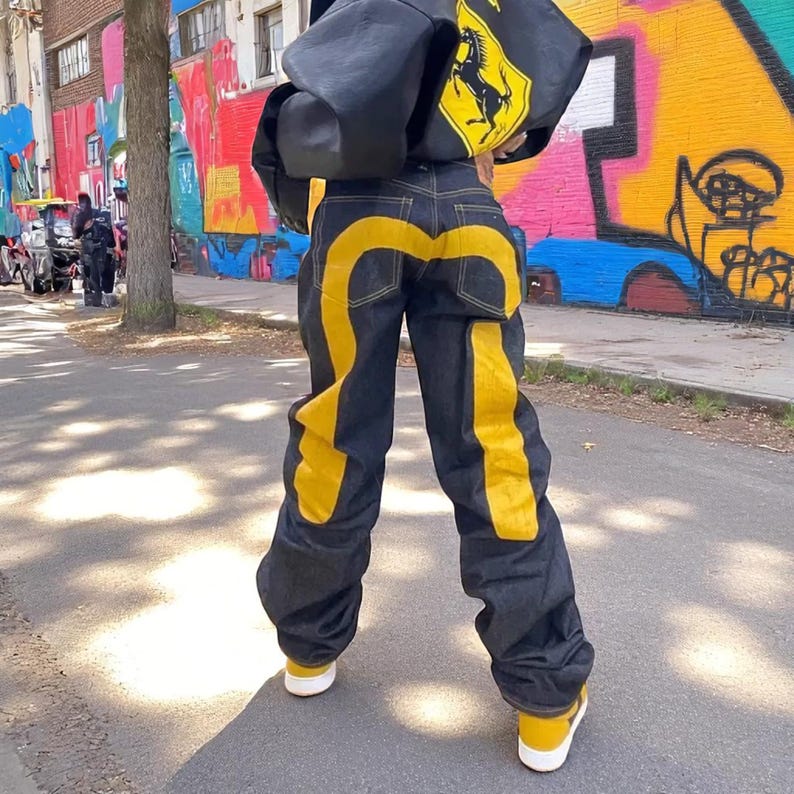 Puede incluir: Vaqueros de mezclilla oscura con llamativos detalles amarillos en los laterales y en la parte trasera. La persona lleva una chaqueta negra con un logotipo amarillo y zapatillas amarillas.