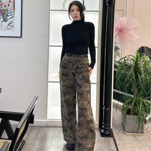 May include: A person wearing a black long-sleeve turtleneck shirt and camouflage print wide-leg pants. The pants are in shades of brown, green, and tan. A black belt and black shoes complete the outfit.