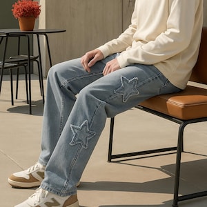 May include: Light blue denim jeans with star-shaped patches on the knees. The jeans are worn with a cream-colored sweatshirt and white and tan sneakers. The person is sitting on a brown leather chair.