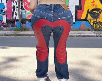 May include: Dark wash denim jeans with a large red heart design on the back. The jeans have a classic five-pocket design and are paired with white and red sneakers. The model is standing in front of a colorful graffiti wall.
