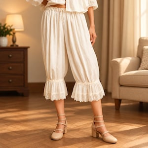 May include: Off-white, calf-length bloomers featuring an elasticized drawstring waistband and gathered fabric at the legs. The hem of each leg is finished with a delicate, scalloped lace trim, creating a soft, layered appearance in the fabric.