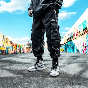 May include: A person wearing black cargo pants with multiple pockets and silver accents. They are wearing silver and black sneakers.
