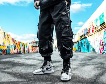 May include: A person wearing black cargo pants with multiple pockets and silver accents. They are wearing silver and black sneakers.