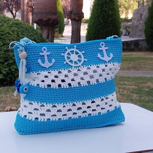 May include: A blue and white crocheted bag with a nautical theme. The bag features white anchors and a white steering wheel. The bag has a blue and white striped pattern and a white crocheted trim.