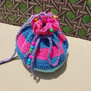May include: A handmade crochet drawstring bag in shades of pink and blue. The bag features horizontal stripes and is adorned with colorful beads. The drawstring and strap are also crocheted, creating a cohesive and vibrant accessory.