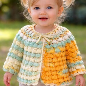 Op de afbeelding: Handgemaakte gehaakte jas in een bubbelsteekpatroon. De jas heeft een colorblock-ontwerp met gele, witte en lichtblauwe secties. Een kleine strik is vastgemaakt bij de halslijn. De jas wordt gedragen door een jong kind.