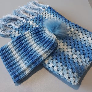 May include: A blue and white crocheted baby hat and blanket set with a fluffy pom pom on the hat.