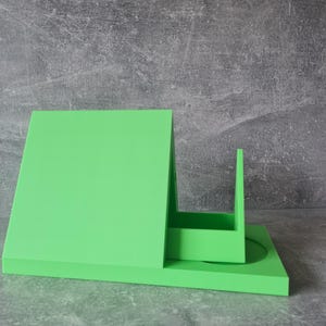 May include: A bright green, geometric desk organizer with a rectangular base, a tall, angled back, and a small, upright compartment. The organizer is made of a solid, smooth material and sits on a gray, textured surface.