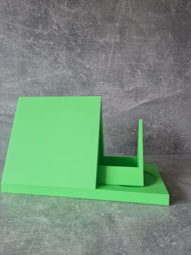 May include: A bright green, geometric desk organizer with a rectangular base, a tall, angled back, and a small, upright compartment. The organizer is made of a solid, smooth material and sits on a gray, textured surface.