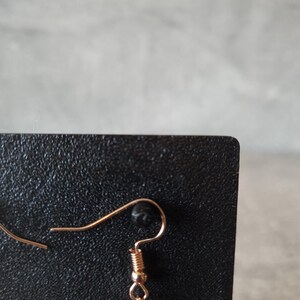 May include: Close-up of a single earring with a gold-colored hook and a red, triangular charm. The charm has black dots and is attached to the hook with a small gold-colored ring. The earring is displayed against a black background.