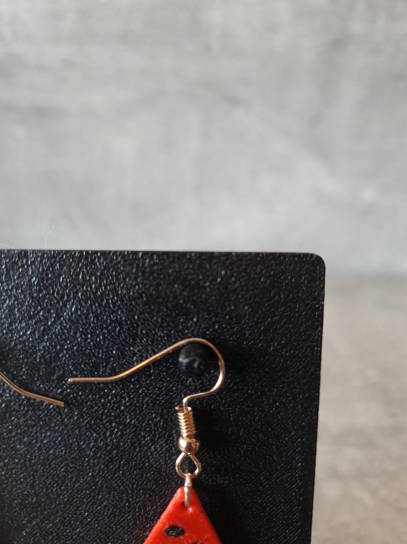 May include: Close-up of a single earring with a gold-colored hook and a red, triangular charm. The charm has black dots and is attached to the hook with a small gold-colored ring. The earring is displayed against a black background.