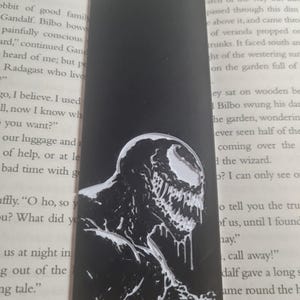May include: Black and white bookmark featuring a detailed illustration of Venom, a fictional superhero character. The bookmark is made of a black material with a white design.