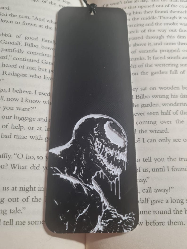 May include: Black and white bookmark featuring a detailed illustration of Venom, a fictional superhero character. The bookmark is made of a black material with a white design.