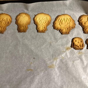 May include: Baked cookies in the shape of characters from a popular fantasy series, including a round-faced owl. The cookies are golden brown and sit on a sheet of parchment paper.