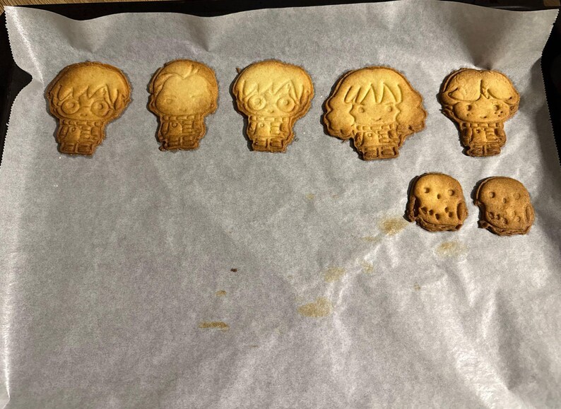 May include: Baked cookies in the shape of characters from a popular fantasy series, including a round-faced owl. The cookies are golden brown and sit on a sheet of parchment paper.