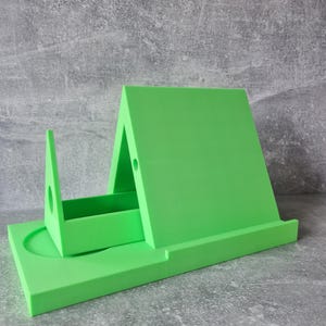 May include: A bright green, 3D-printed phone stand. The stand has a rectangular base with a circular indentation, a vertical support, and an angled back. The stand is designed to hold a phone or tablet. The stand measures approximately 6 inches long.