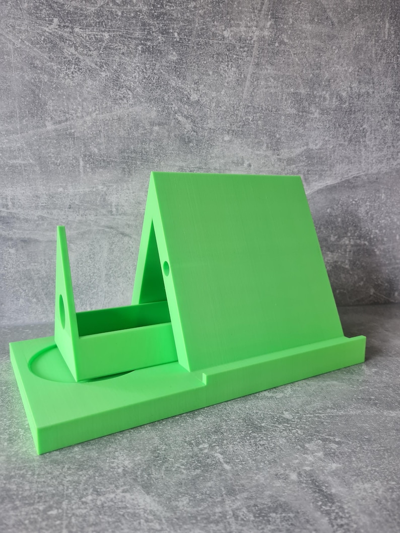 May include: A bright green, 3D-printed phone stand. The stand has a rectangular base with a circular indentation, a vertical support, and an angled back. The stand is designed to hold a phone or tablet. The stand measures approximately 6 inches long.