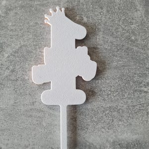 Op de afbeelding: Een witte taarttopper in de vorm van een giraffe met een kroon, met het nummer "1" in het ontwerp verwerkt. De topper heeft een getextureerd oppervlak en een puntige stok om in een taart te steken.