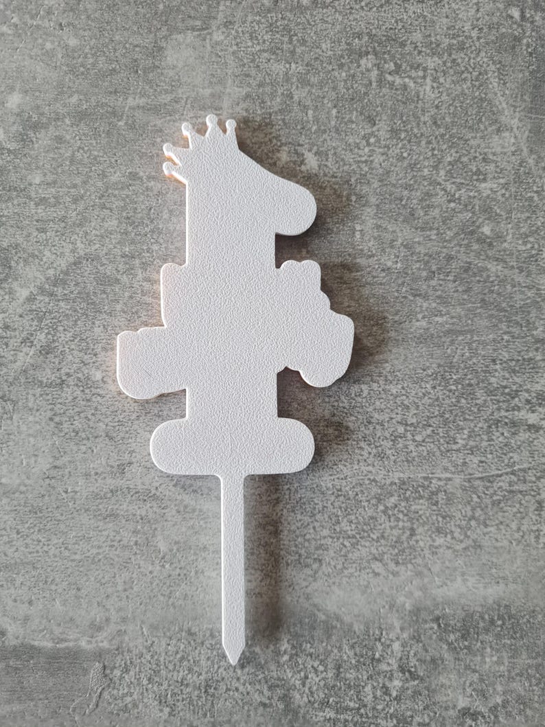 Op de afbeelding: Een witte taarttopper in de vorm van een giraffe met een kroon, met het nummer "1" in het ontwerp verwerkt. De topper heeft een getextureerd oppervlak en een puntige stok om in een taart te steken.