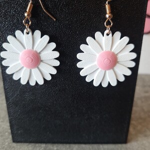 May include: A pair of daisy earrings. Each earring features a white daisy with a pink center. The daisy petals are detailed. The earrings have gold-colored hooks.